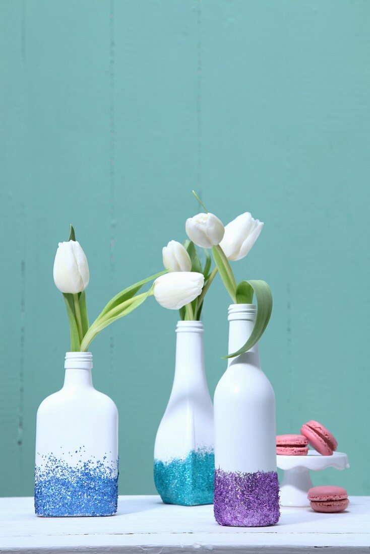 Weiß bemalte Flaschen mit buntem Glitzer als Vasen mit weissen Tulpen vor türkisfarbener Wand