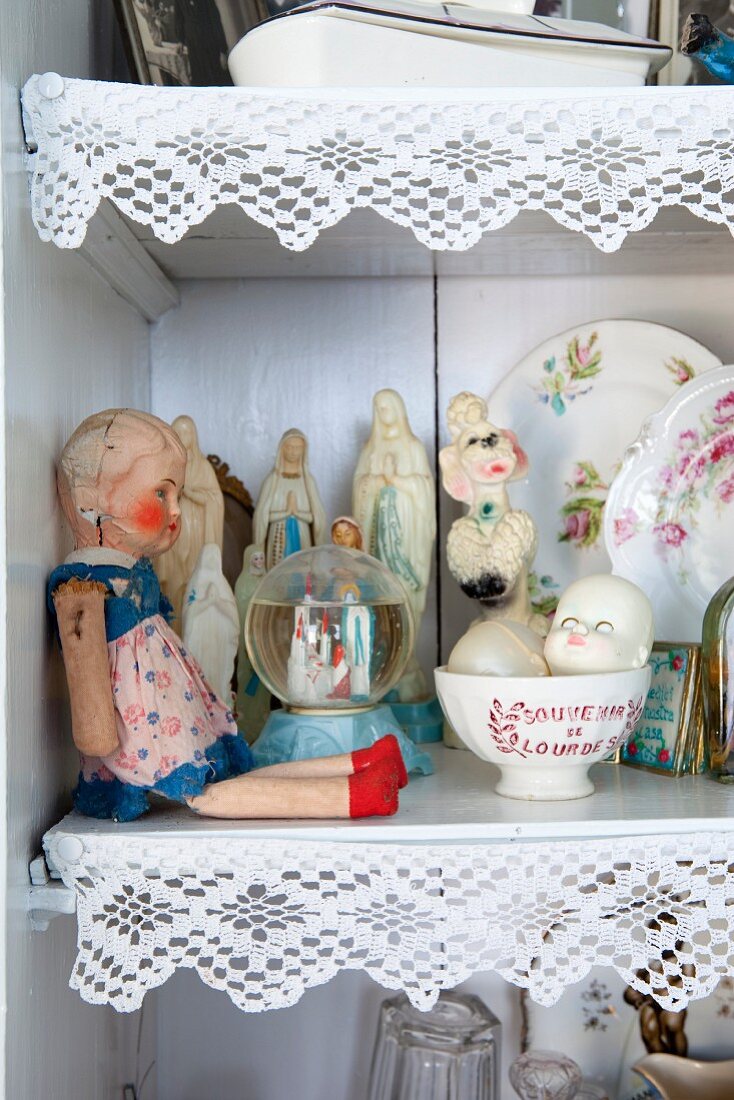 Puppe und Sammlerstücke vor Devotionalien im Regalschrank, Ablagen mit weisser Borte verziert