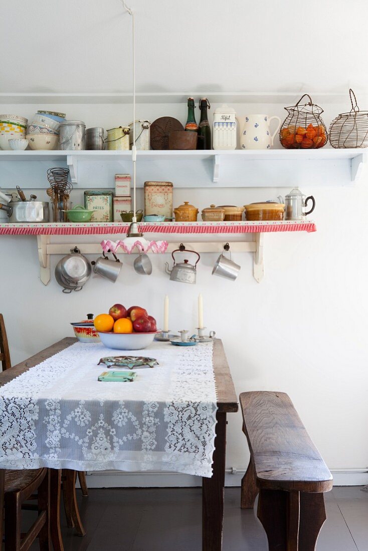 Lace tablecloth on dining table, antique bench and retro crockery on wall-mounted shelves