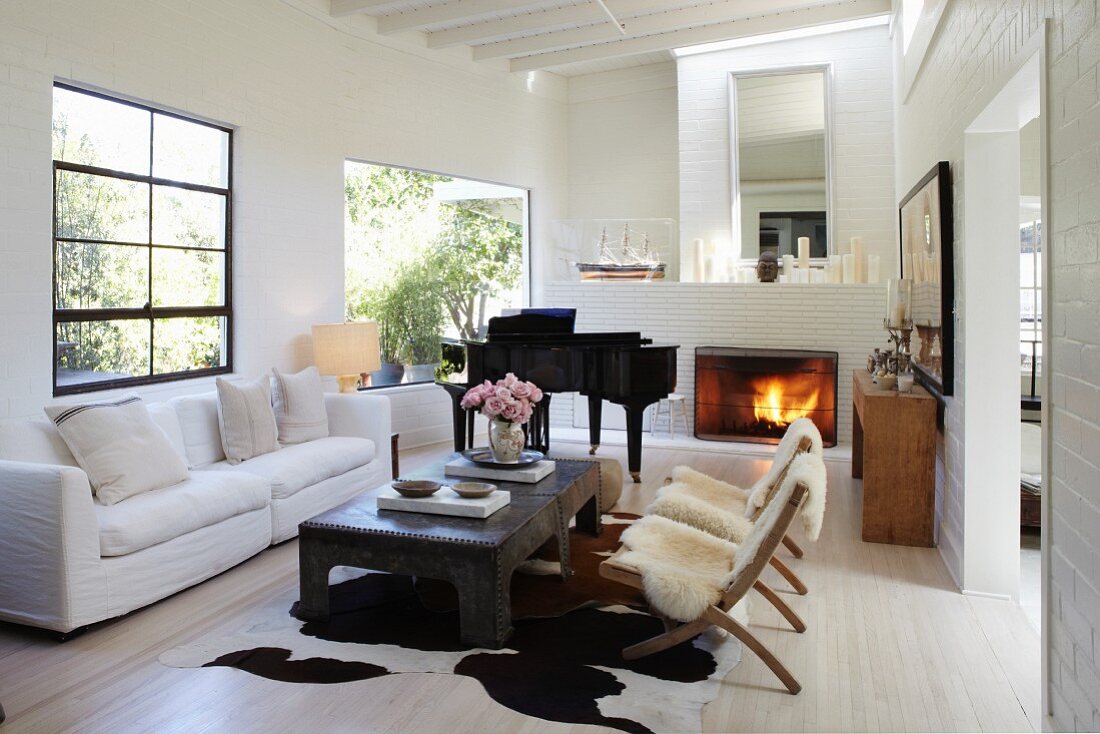 Chairs with sheepskin blankets and white sofa around coffee table; grand piano next to open fireplace in background in living room