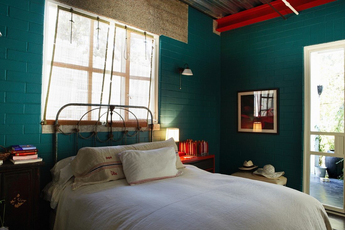 Bed with vintage metal frame below window and table lamp on red bedside cabinet in simple bedroom with brick walls painted dark green