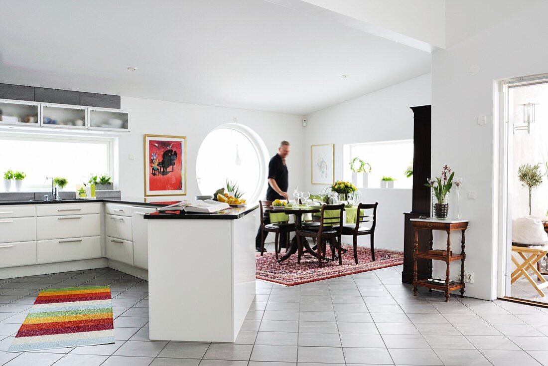White, open-plan kitchen with expansive counter and man in traditional-style dining area to one side in open-plan interior