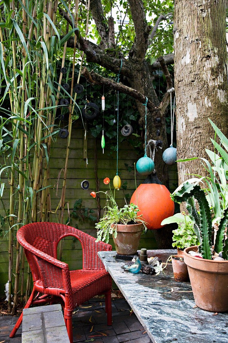Red-painted wicker armchairs and potted plants on stone counter in small courtyard, colourful pedants hanging from branch in background