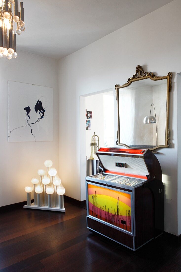 Retro jukebox under gilt-framed, antique mirror, modern artwork and unusual light sculpture in white interior