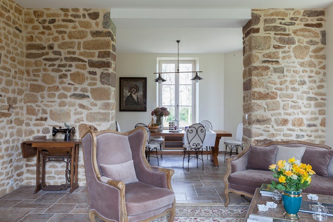 Sofa and armchair in modernised, rustic interior with stone wall and view of dining area through floor-to-ceiling open doorway