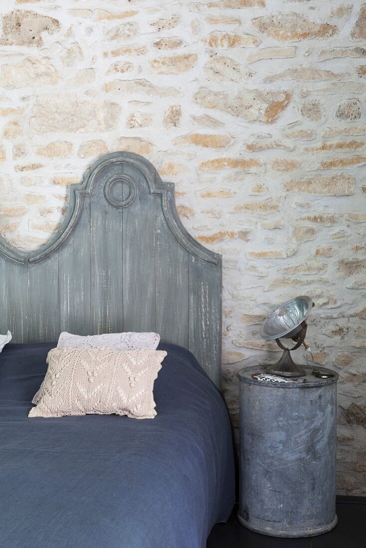 Grau gestrichenes Bettkopfteil an rustikaler Natursteinwand, Blechtonne als Nachttisch mit Strahler