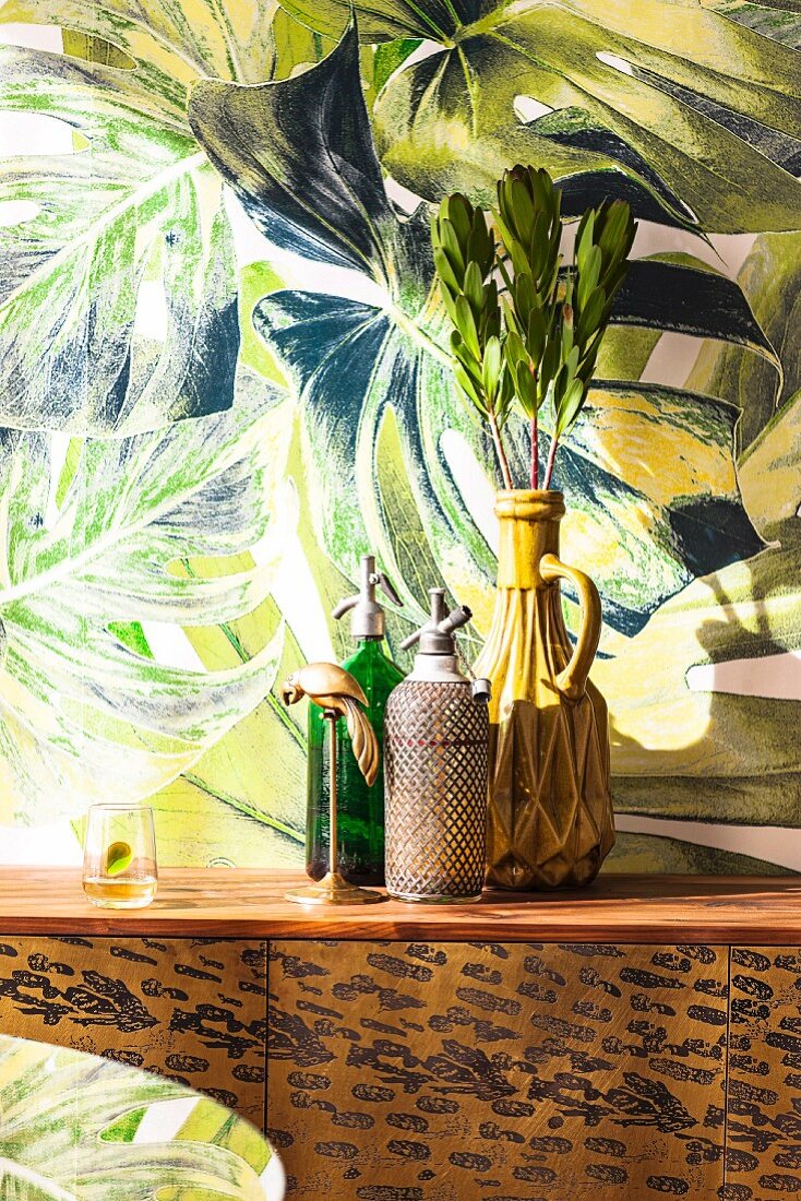 Stillleben aus Retro Sodaflaschen und Vase mit tropischen Bätterzweigen auf Sideboard, vor Wand mit expressiver Malerei