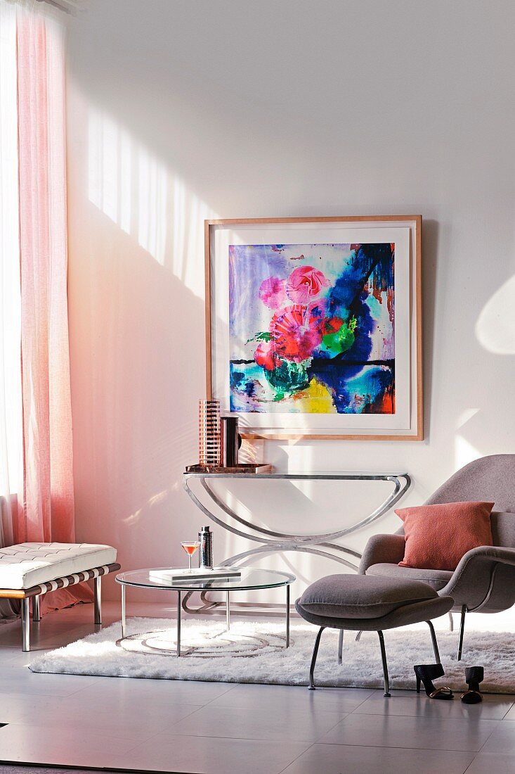 Pale grey armchair and footstool next to round coffee table on white flokati rug in front of console table below floral painting