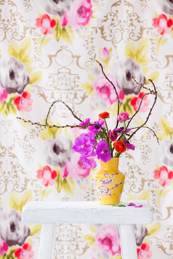 Anemonen, Ranunkeln und Orchidee in gelb geblümter Vase vor floral gemusterter Tapete