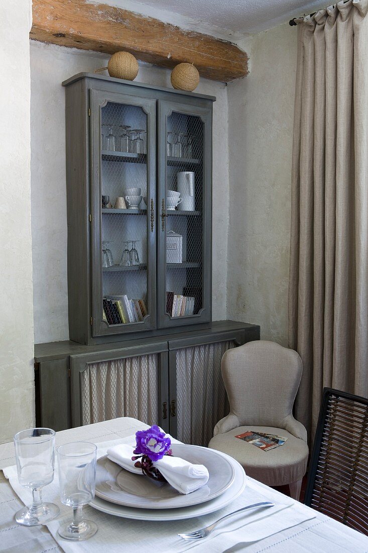 Wooden beam above grey-painted crockery cabinet with fabric panels in lower doors, upholstered chair and set table in foreground