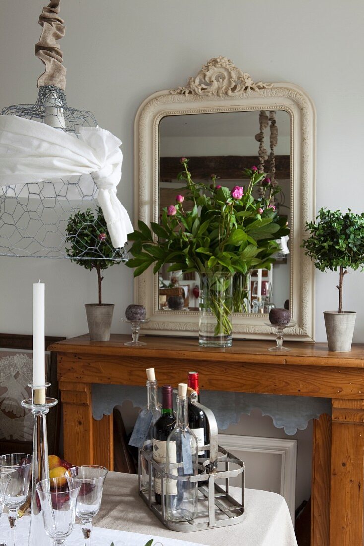 Konsolentisch mit antikem Spiegel und Pfingstrosenstrauß, Vintage Flaschenträger mit Weinflaschen auf gedecktem Tisch
