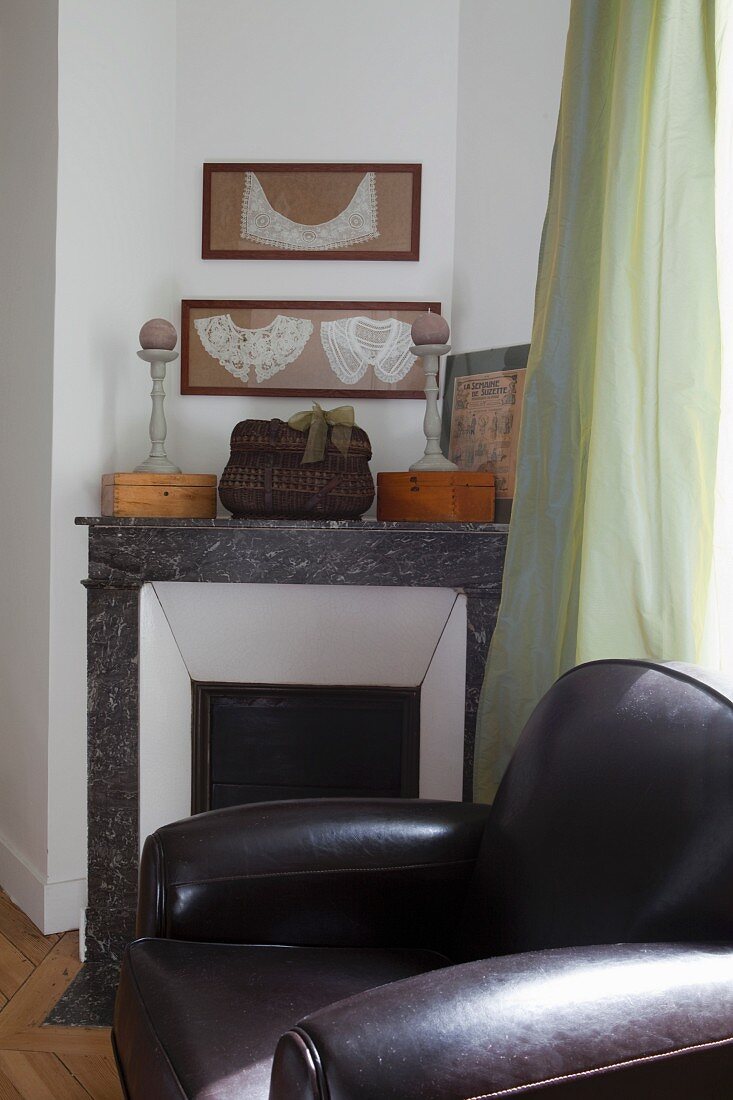 Black leather armchair in front of disused fireplace with ornaments on marble surround below framed, antique lace collars