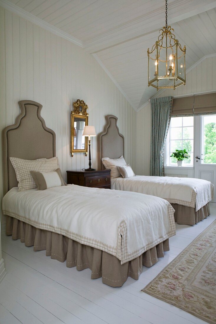Zwei Einzelbetten mit gepolstertem Kopfteil, Messingleuchte und goldgerahmter Spiegel in holzverkleidetem Schlafzimmer