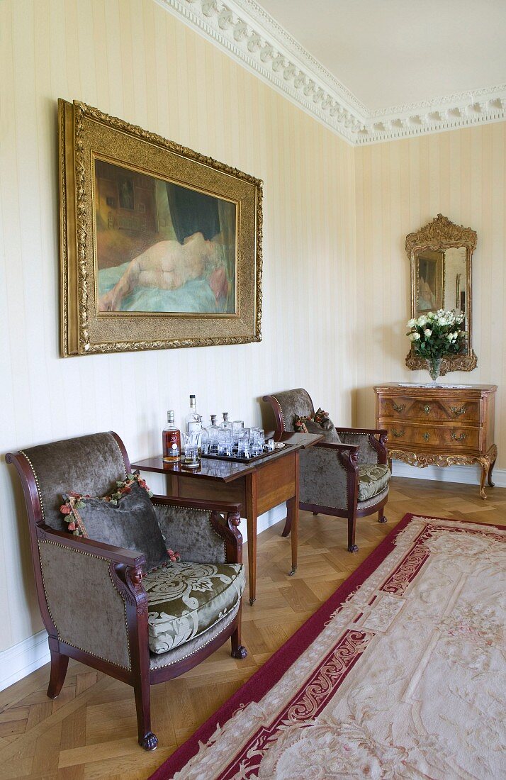 Antikstühle und Tisch unter Ölgemälde mit Frauenakt, zart gestreifte Tapetenwand und Barockkommode mit Spiegel im Hintergrund