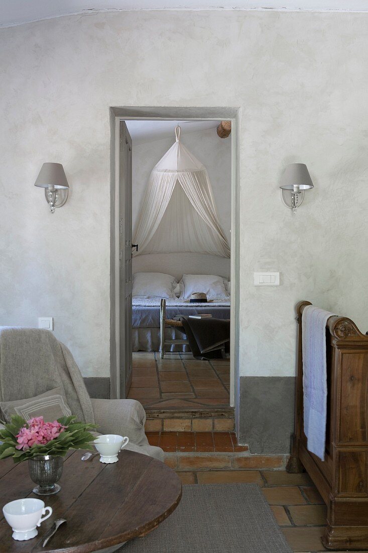 Holztisch mit nostalischen Kaffeetassen, Sessel und Recamiere, Blick durch offene Tür auf Doppelbett mit Moskitonetz