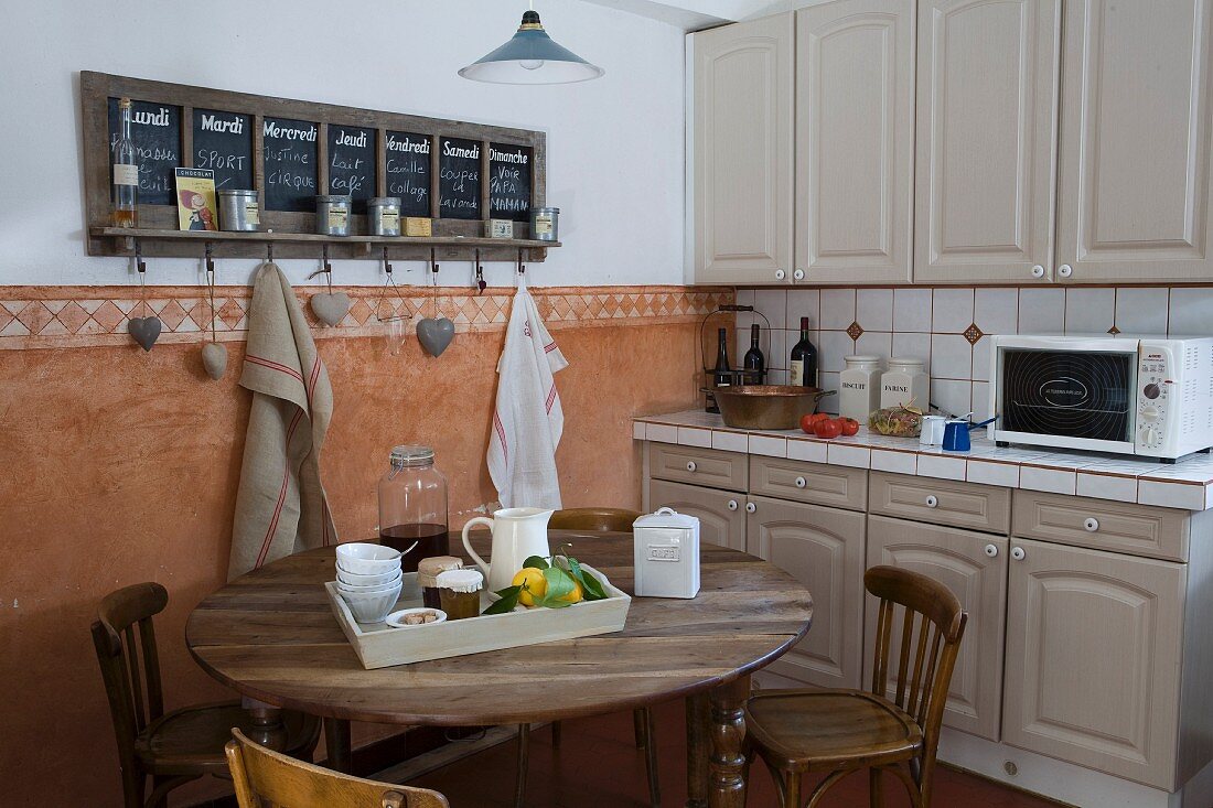 Mediterranean country-house kitchen counter with grey cupboards and white tiles, dining table, wooden chairs, and chalkboard calender above terracotta dado