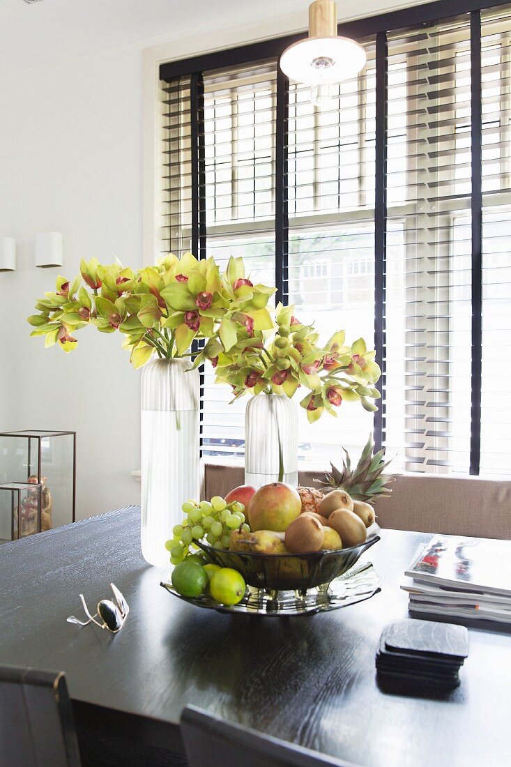 Obstschale und Vasen mit Blumenzweigen auf schwarzem Esstisch, im Hintergrund dunkle Jalousie am Fenster
