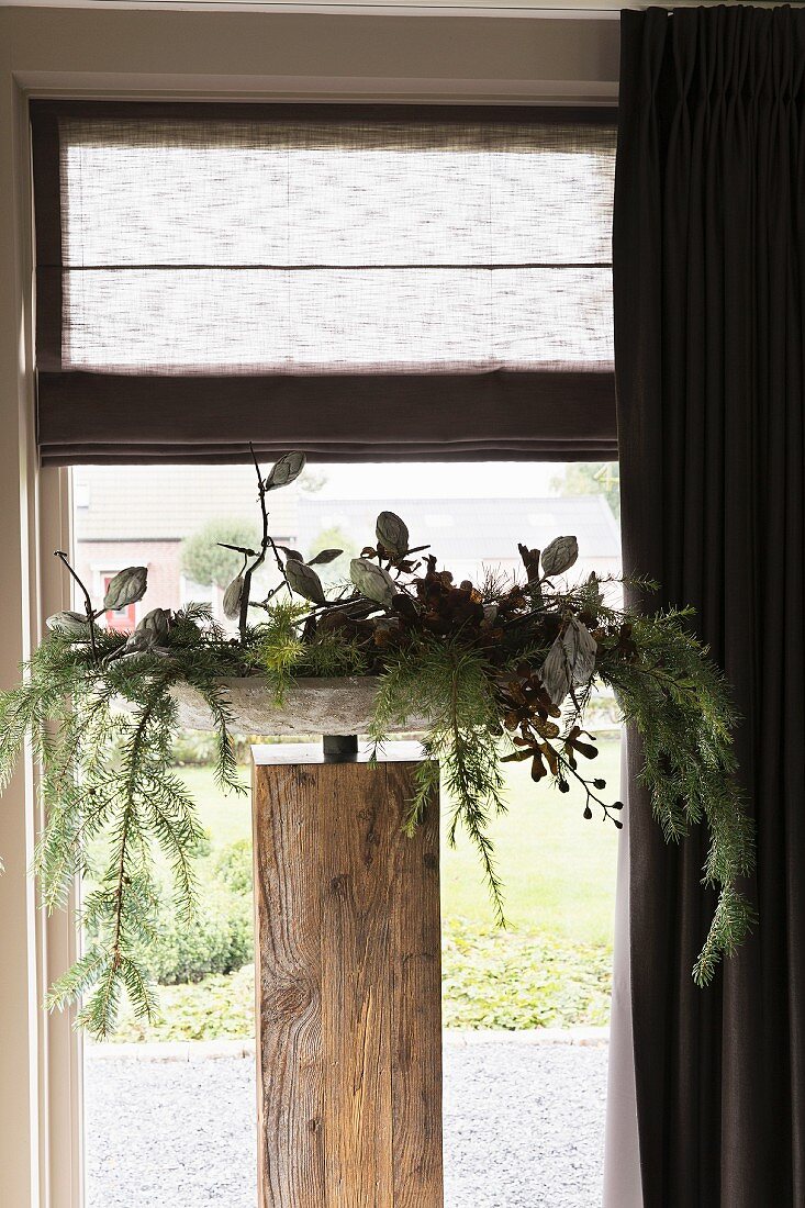 Weihnachtlich dekorierte Schale auf Holzstele, vor Fenster mit Raffrollo