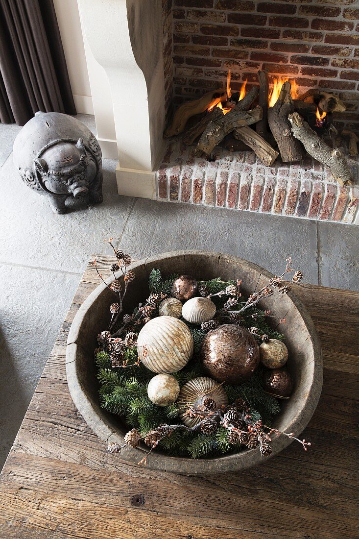 Christmas arrangement in wooden bowl on rustic table and pig ornament next to open fire in fireplace
