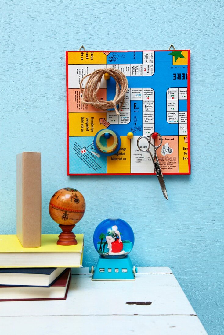 Retro-Brettspiel als Pinnwand mit Aufhängungen aus Spielfiguren für Schere, Schnur und Klebeband über Schreibtisch mit Bücherstapel