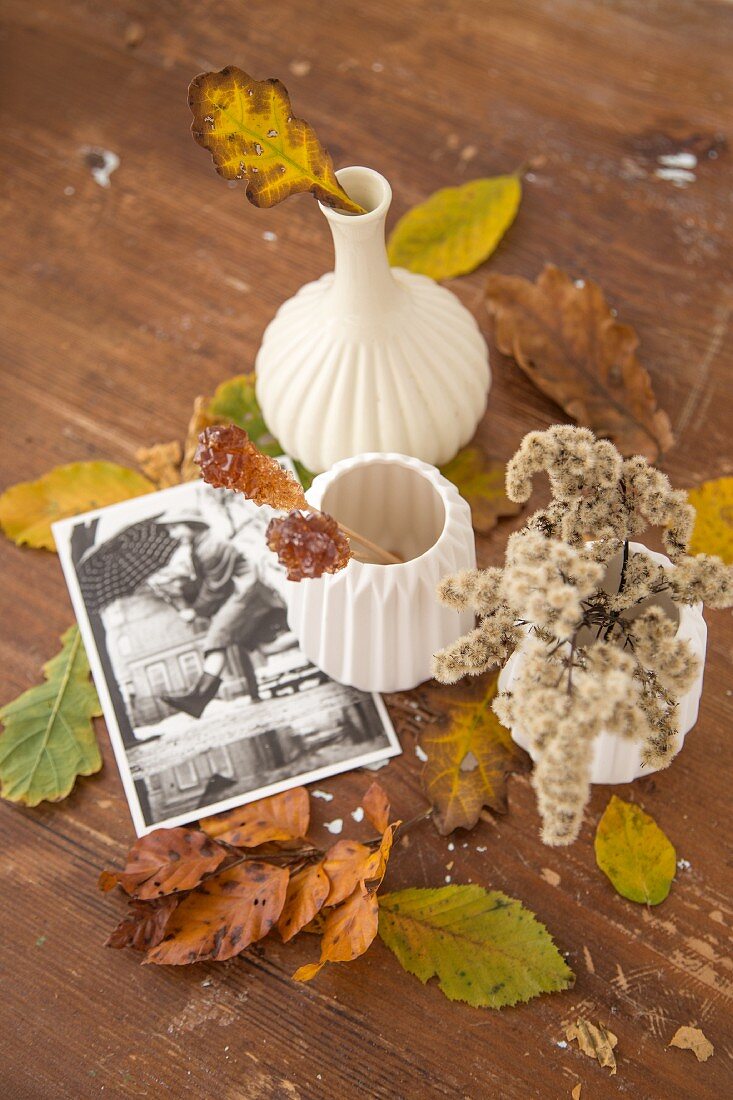 Verfärbte Blätterzweige in weissen Keramikvasen zwischen Herbstlaub und Postkarte auf Holzuntergrund