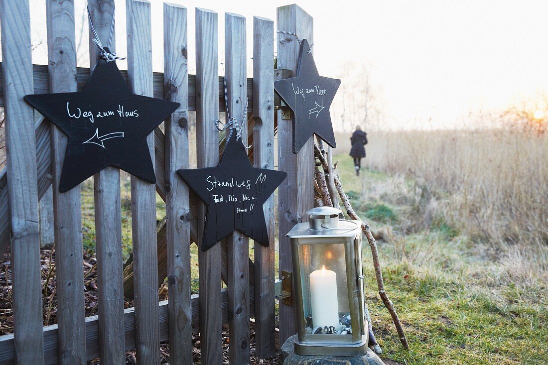 Sternförmige Wegweiser aus schwarz lackiertem Holz an Zaun gehängt, davor Laterne mit Kerze, im Hintergrund Spaziergängerin