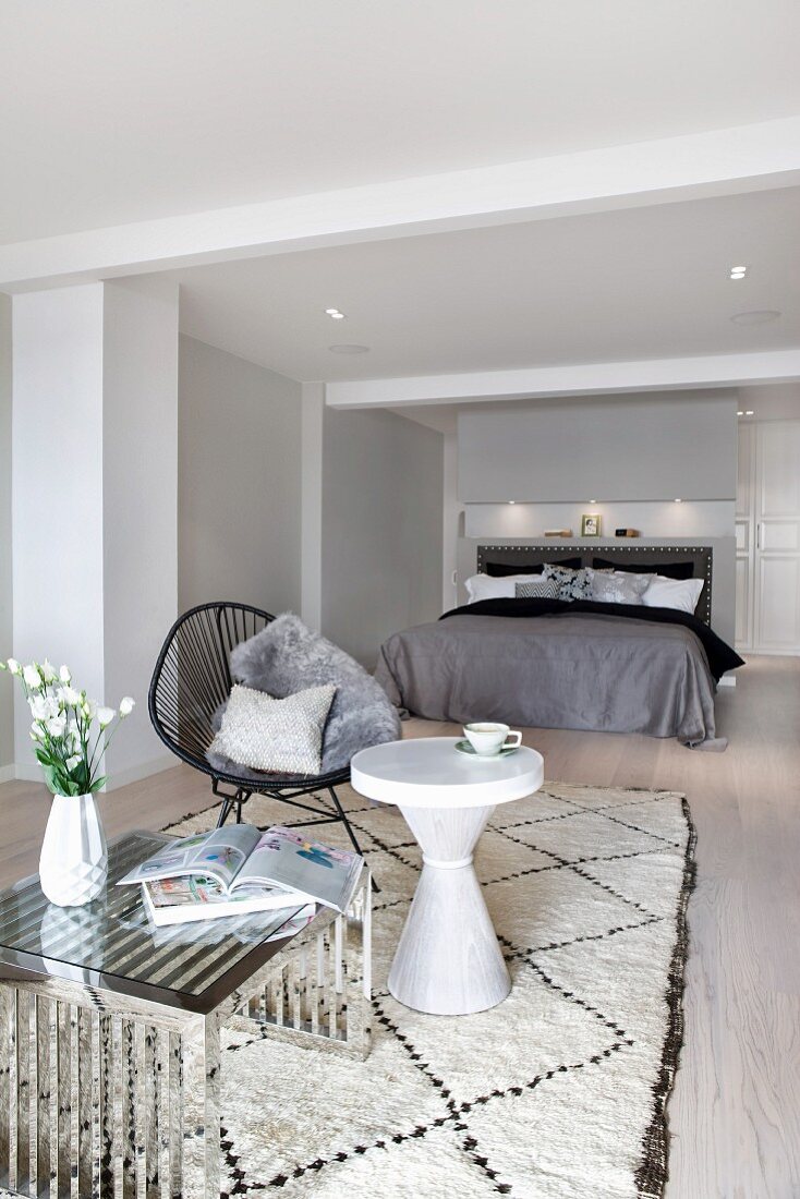 Chrome-framed coffee table, white side table and black easy chair on rug in front of double bed in minimalist bedroom