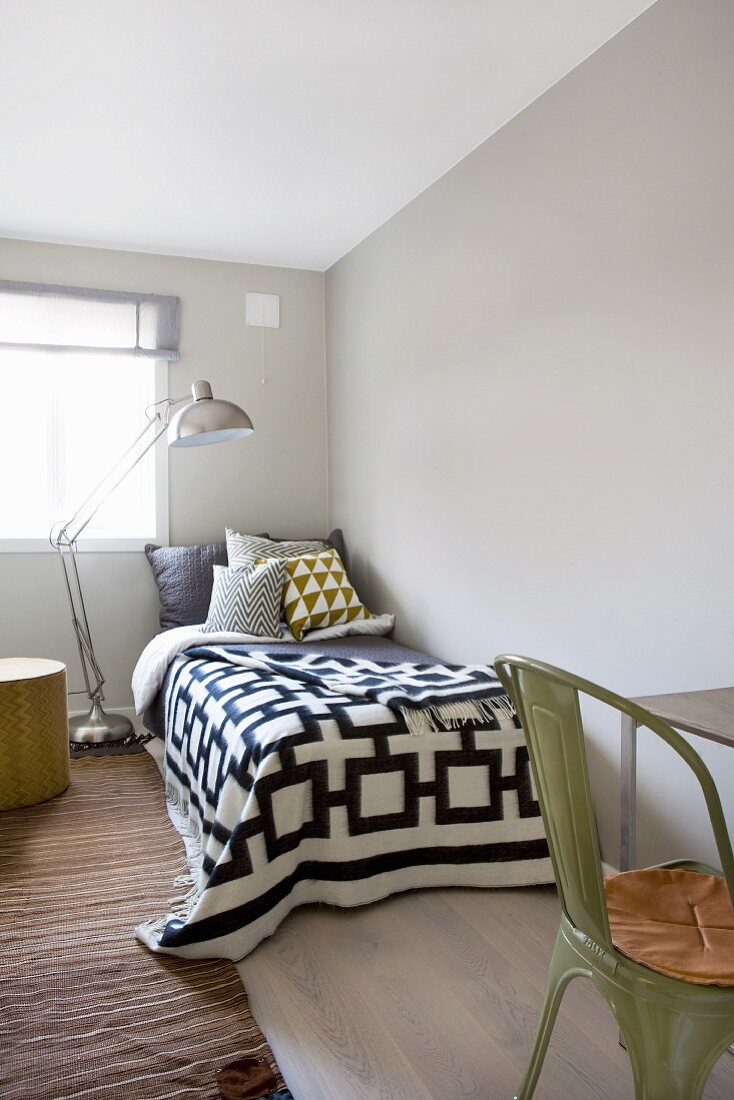 Schwarz-weiße Tagesdecke mit geometrischem Muster auf Bett, daneben Retro Stehleuchte vor Fenster, im Vordergrund grüner Metallstuhl