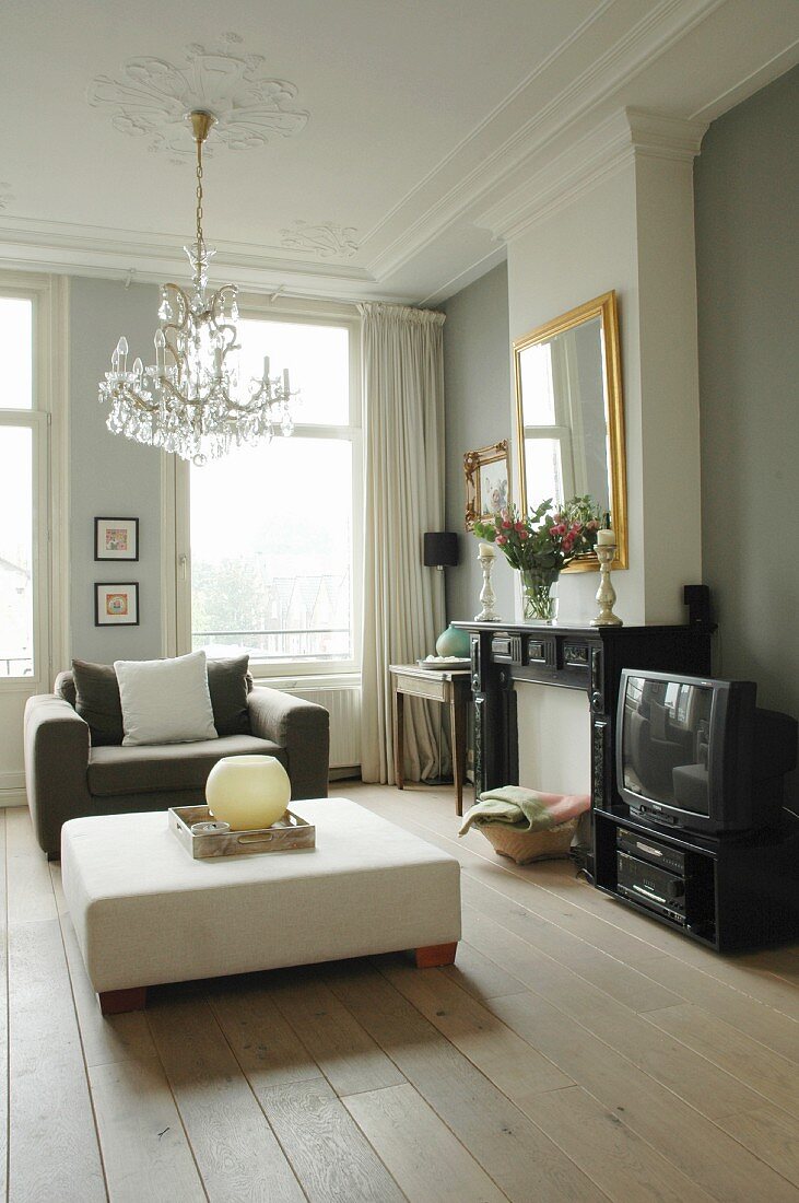 Herrschaftliches Wohnzimmer mit minimalistischem Flair, weisser, gepolsterter Bodentisch und grauer Polstersessel unter Kronleuchter an Stuckdecke