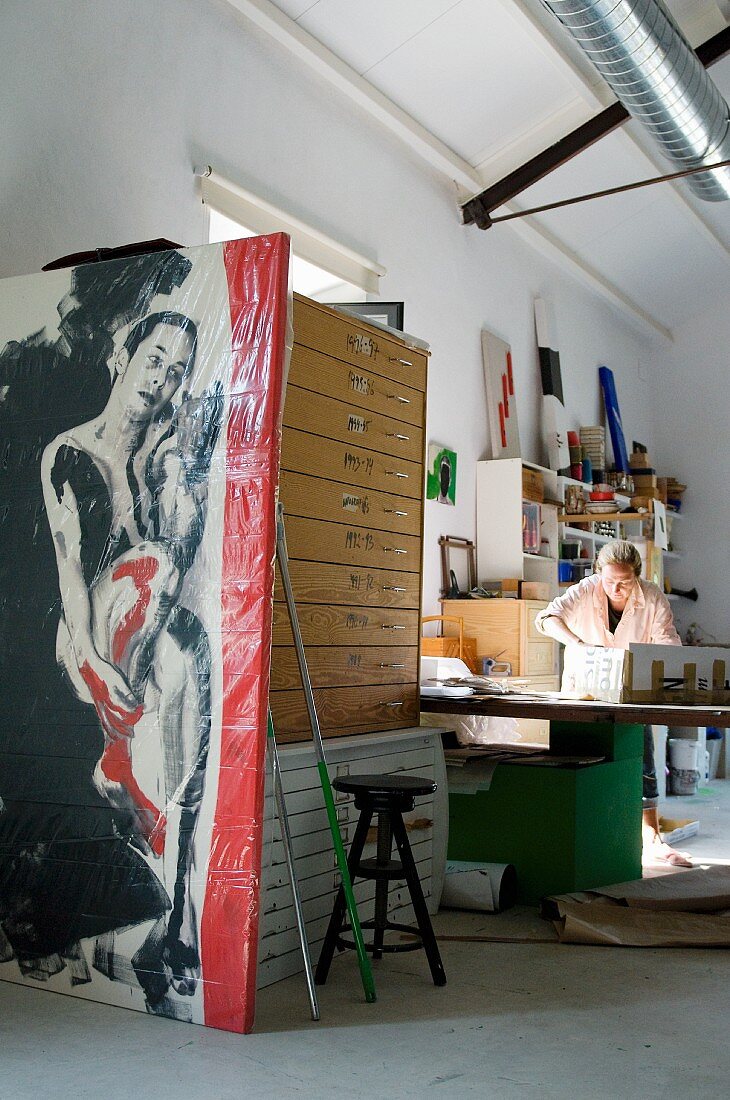 Atelierausschnitt - Frau am Arbeitstisch, im Vordergrund verpacktes Bild vor Planschränken