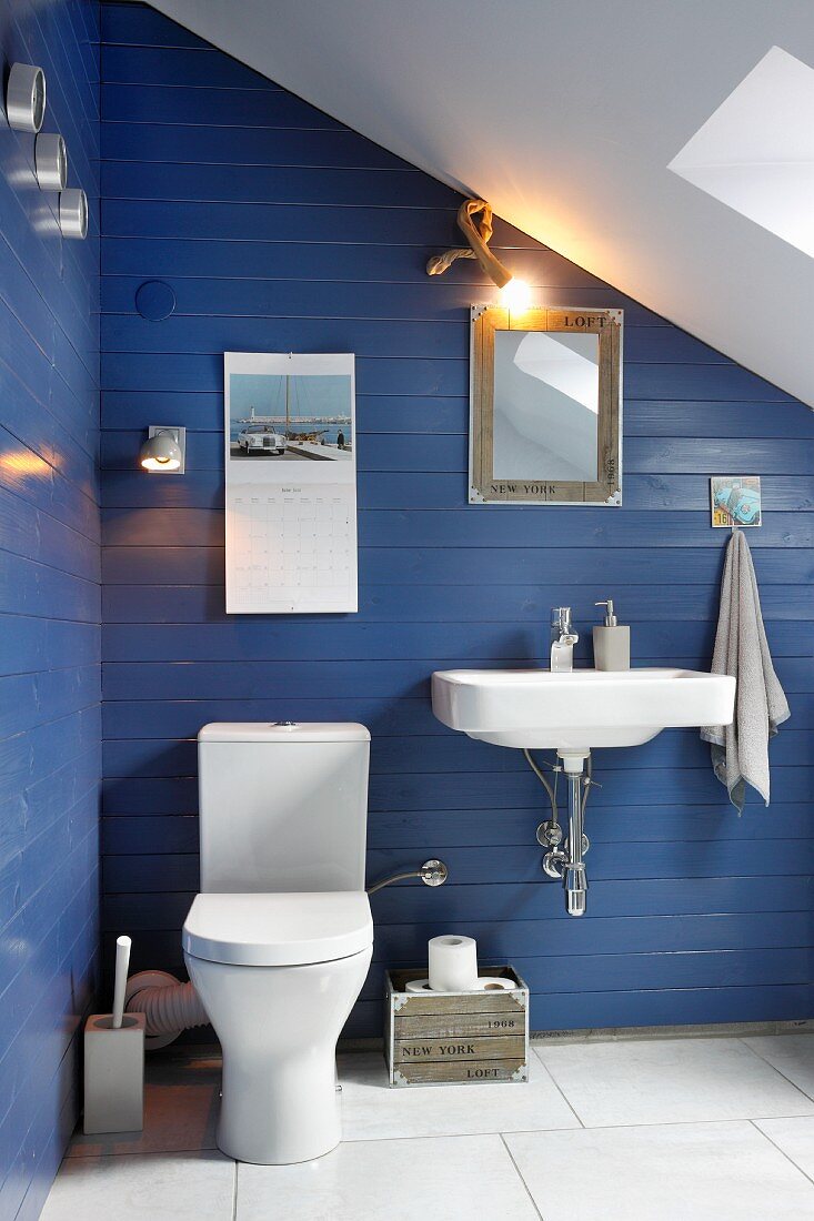 Weisses Waschbecken und Toilette an blau gestrichener Holzwand im Bad unter dem Dach