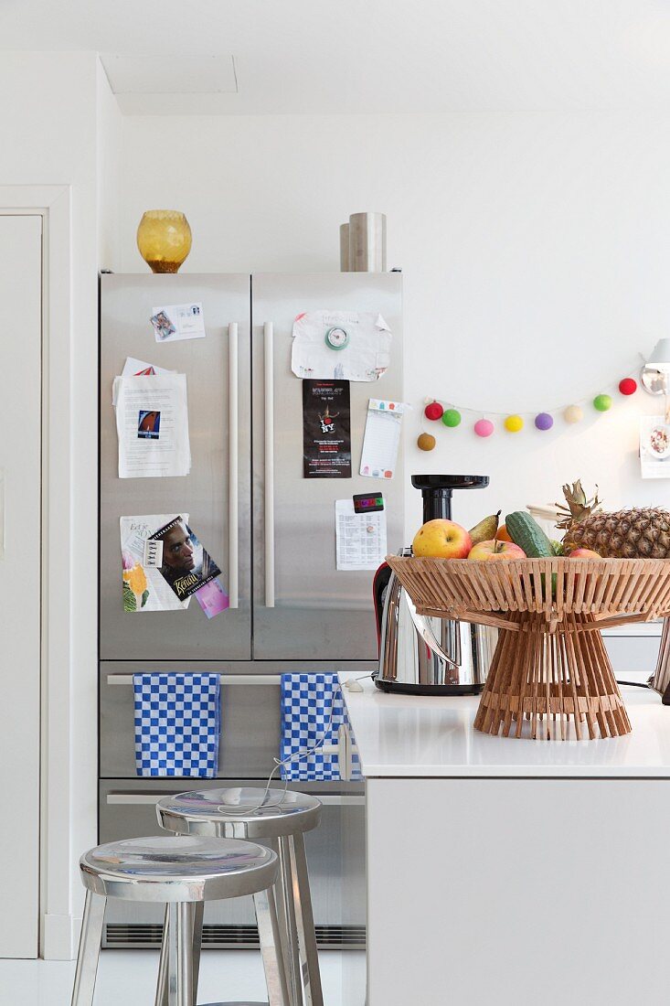 Metall Barhocker vor Theke, darauf Obstkorb, im Hintergrund Edelstahl Kühlschrankkombination in heller Küche