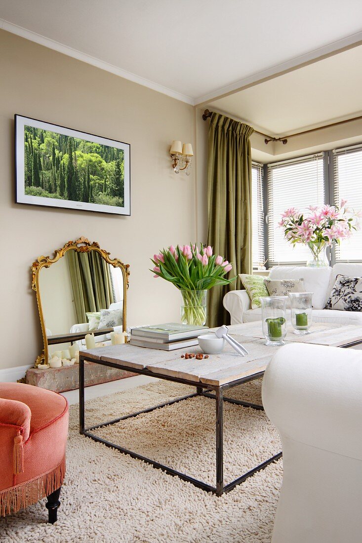 Delicate coffee table with rustic wooden top on long-pile rug, vases of flowers and gilt-framed mirror on wall in classic living room