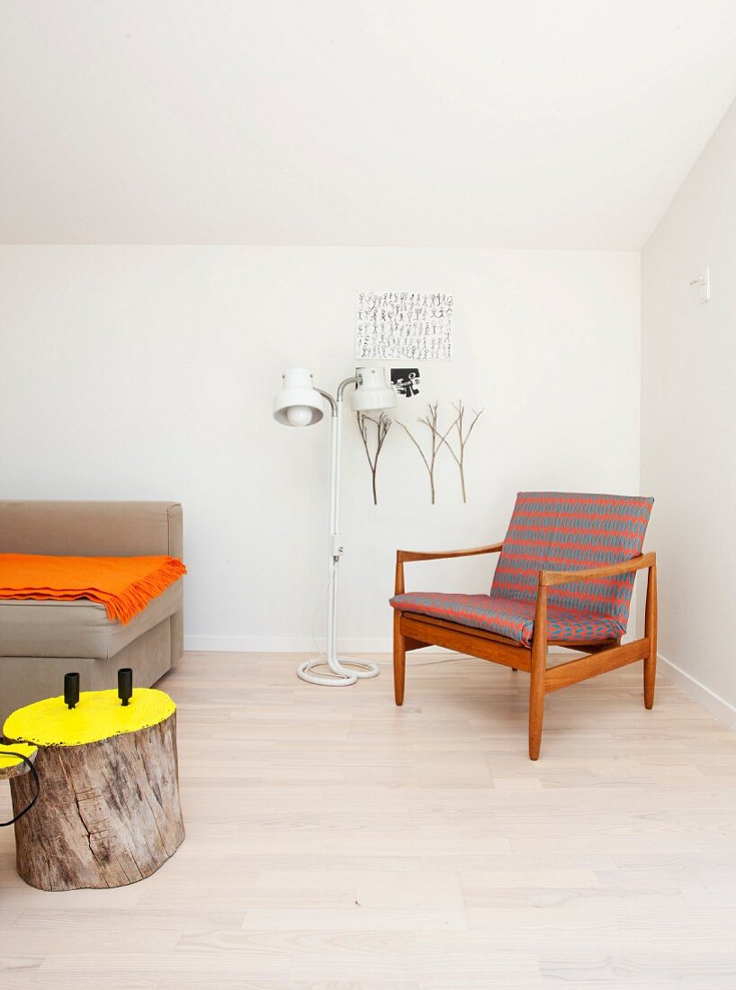 Sessel mit Holzgestell im Fiftiesstil neben Stehleuchte , im Vordergrund Baumstamm als Beistelltisch mit gelb gestrichener Schnittfläche