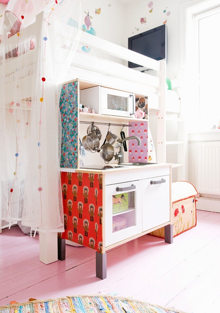 Child's bedroom with tulle curtains on white loft bed, pale wooden floor and patterned wallpaper