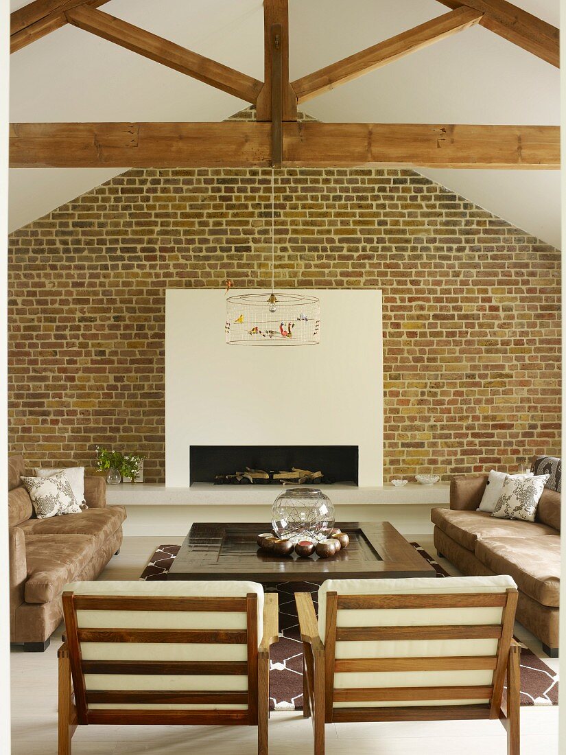 Armchairs with white cushions on wooden frames and sofas around low coffee table in front of fireplace in white chimney breast on brick wall