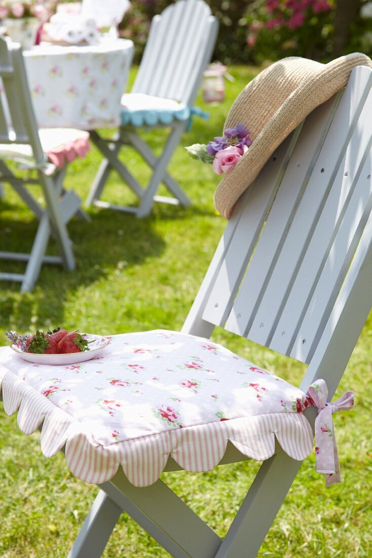 Romantic floral seat cushion on pale grey garden chair decorated with dish of strawberries and sun hat