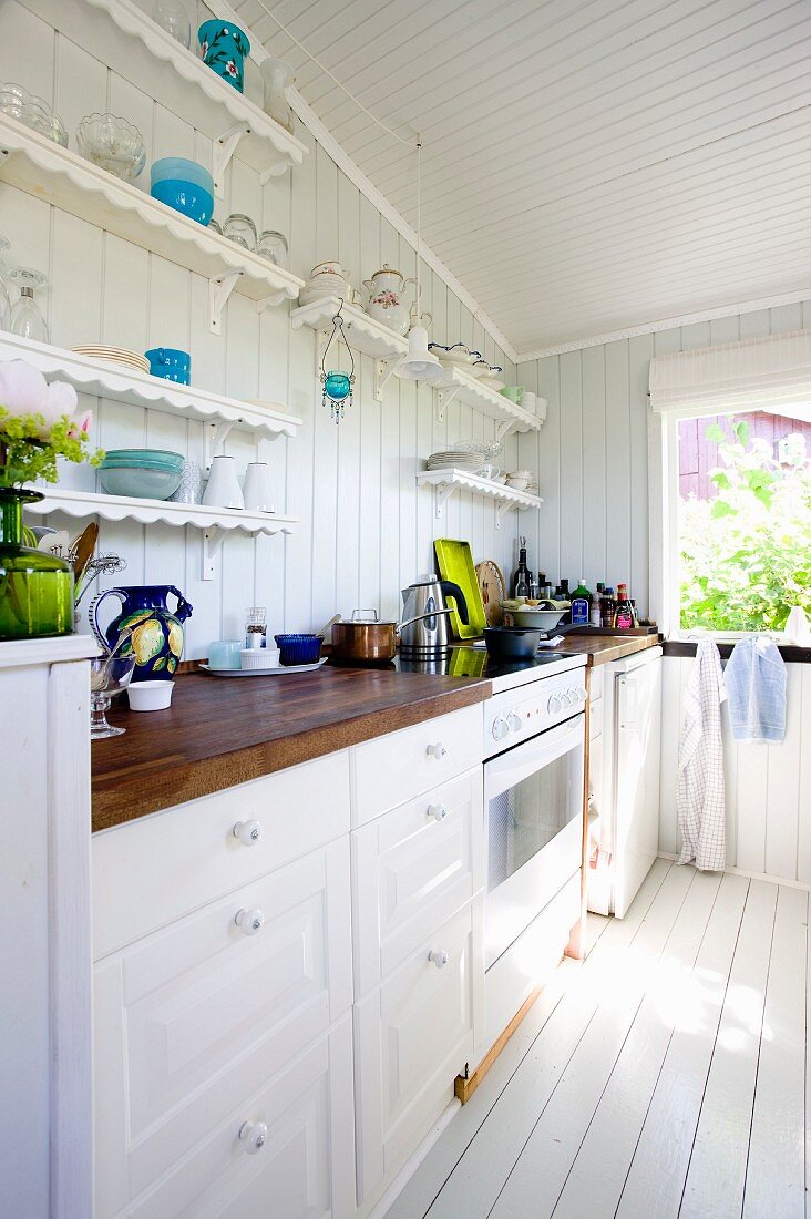 White kitchen counter in traditional country-house style below shelves of ornaments on white wooden wall