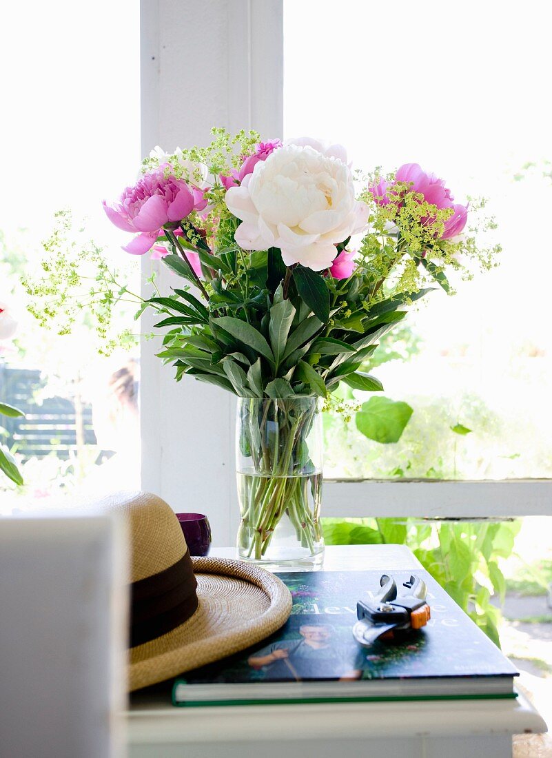 Peonies in glass vase, summer hat, gardening book and secateurs on white table in front of window