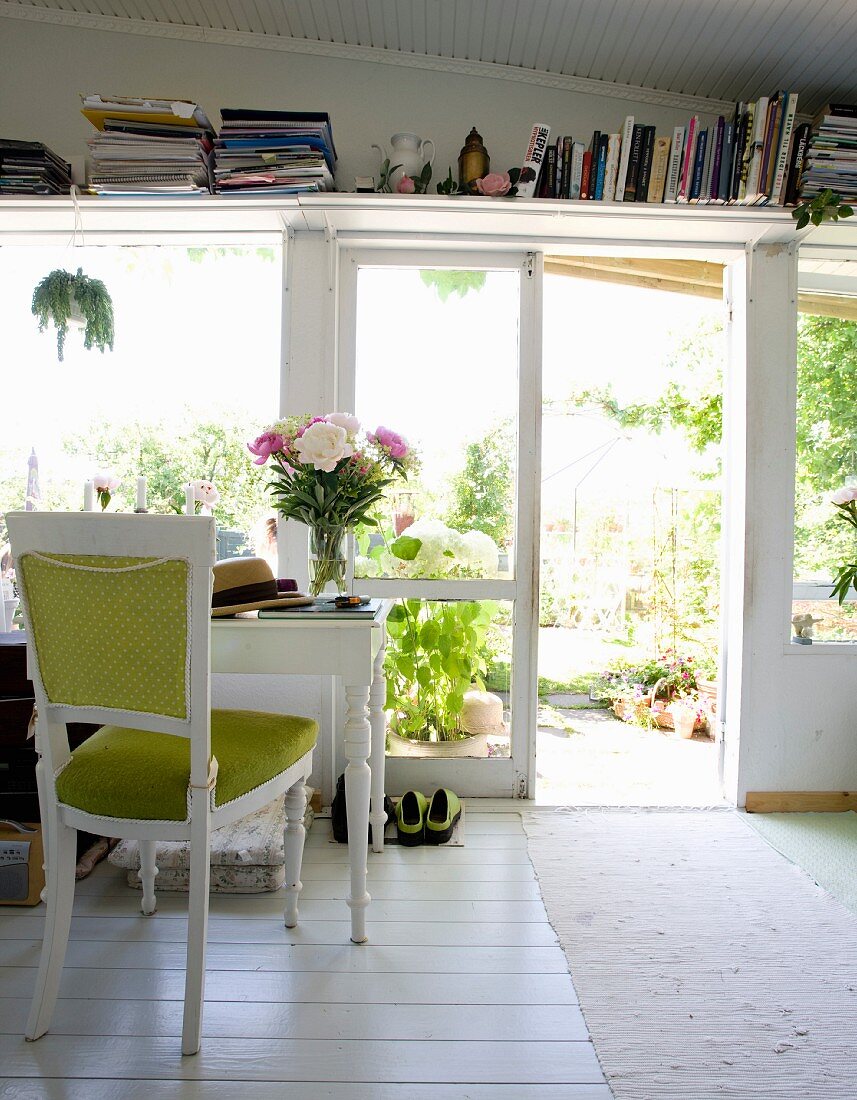 Vollgestelltes Bücherbrett und weißer nostalgischer Schreibtisch mit Polsterstuhl vor offener Terrassentür