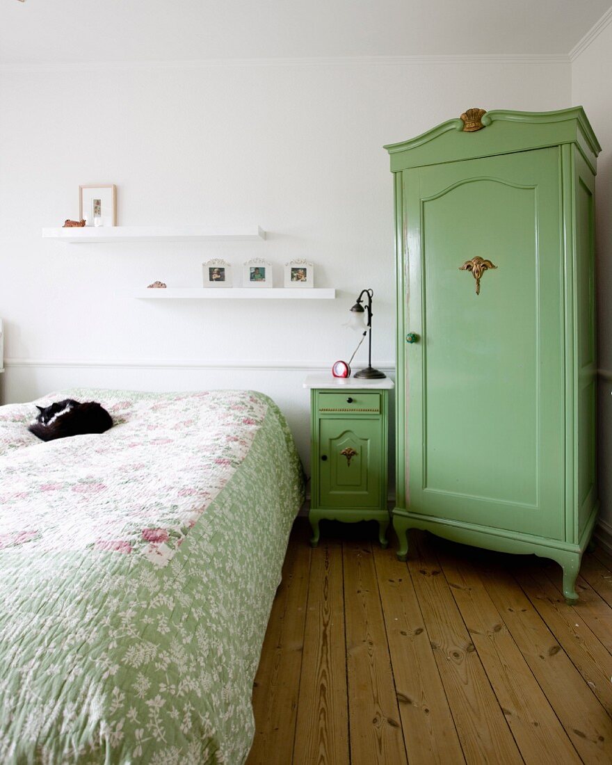 Rustic corner cupboard painted green, bedside cabinet and cat on double bed with patterned bedspread