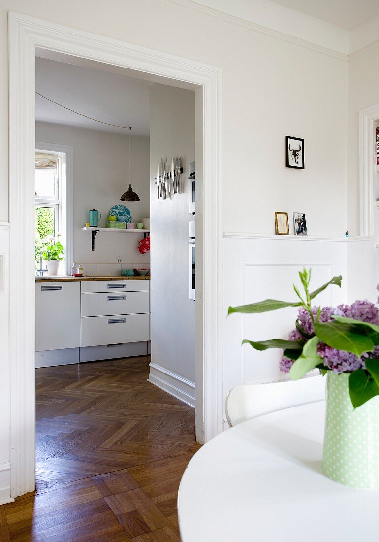 Weißer Tisch mit Blumenvase und Blick durch offenen Türdurchgang in moderne Küche, edler Fischgrätparkett