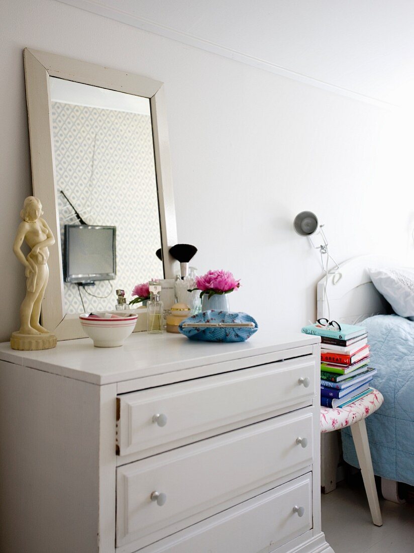 Make-up brushes and framed mirror on chest of drawers next to three-legged stool and bed in bedroom