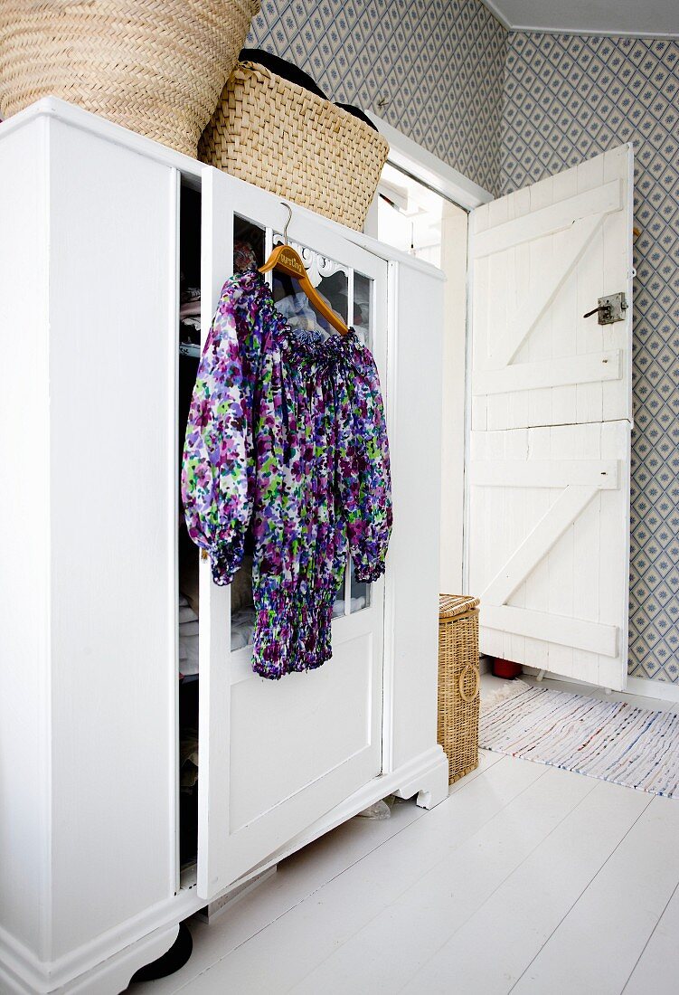 Kleiderbügel mit geblümtem Kleid an weißem Kleiderschrank aufgehängt, im Schlafzimmer mit gemusterter Tapete an Wand