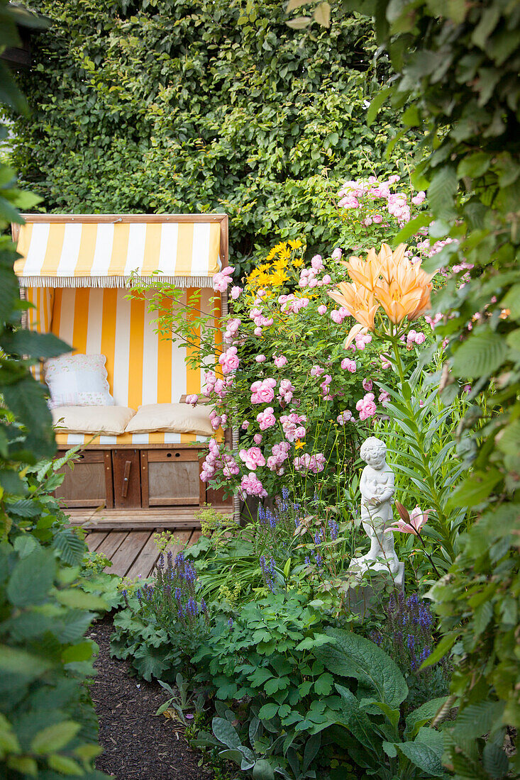 Blühende Lilie und Rosenbusch vor verstecktem Sitzplatz, Strandkorb gelb-weiss gestreift in idyllischem Garten