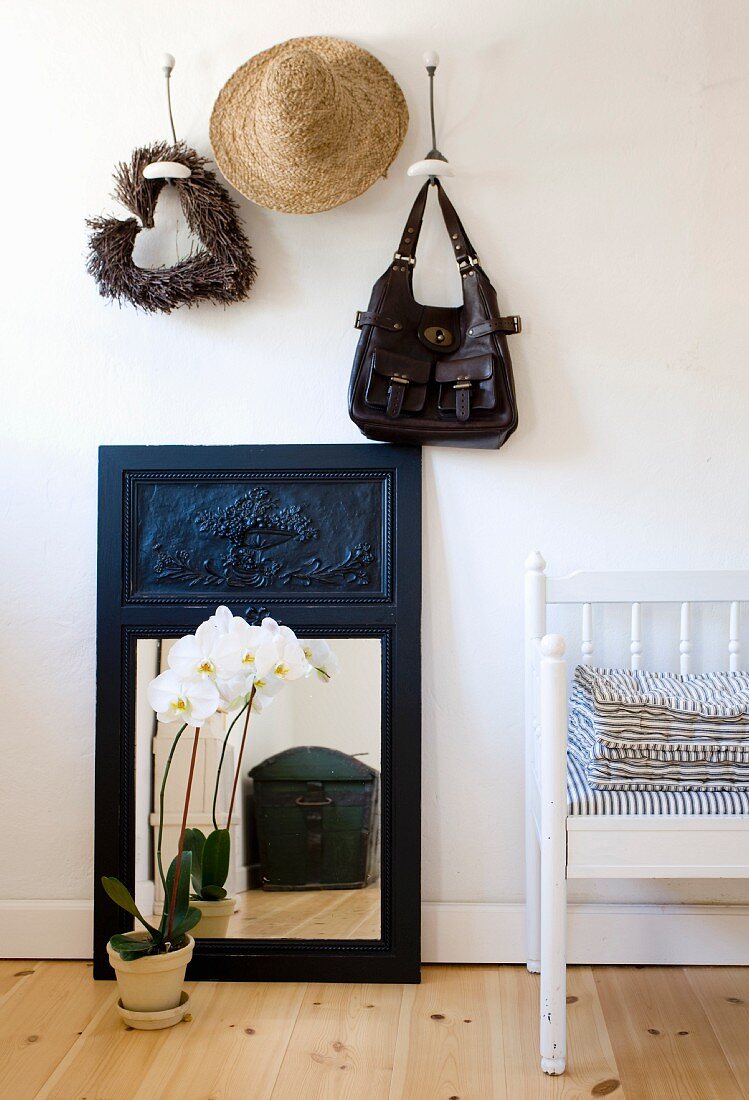 weiße Orchidee im Topf vor Spiegel auf Dielenboden, darüber Garderobe mit aufgehängter Tasche und Hut an Wandhaken, seitlich teilweise sichtbare Sitzbank