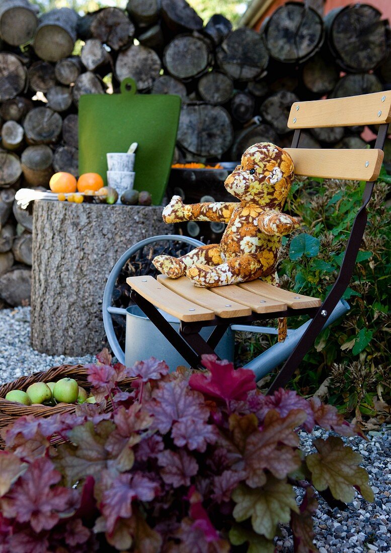 Teddybär aus gemustertem Stoff auf Holz Klappstuhl neben Baumstamm als Tisch im Garten