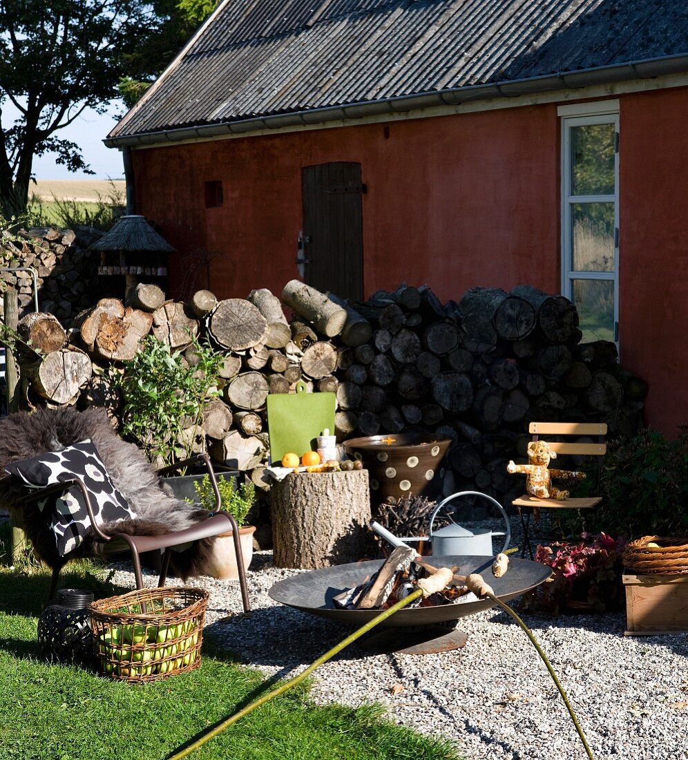 Metal outdoor armchairs and fire bowl on terrace with campfire bread over fire and firewood stacked against house