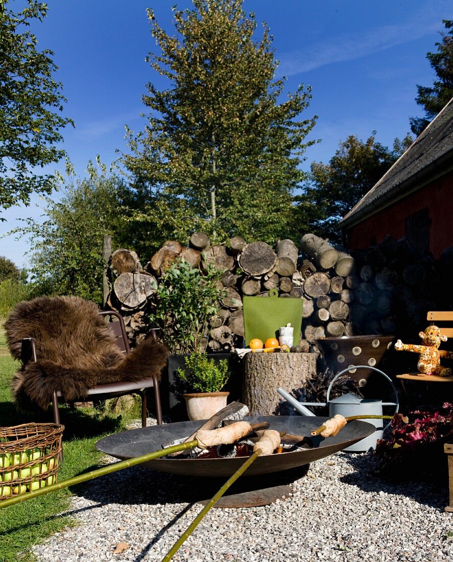 Stockbrot auf Feuerschale, auf gekiestem Platz im Garten, im Hintergrund Outdoor Metallsessel mit braunem Tierfell vor gestapeltem Brennholz