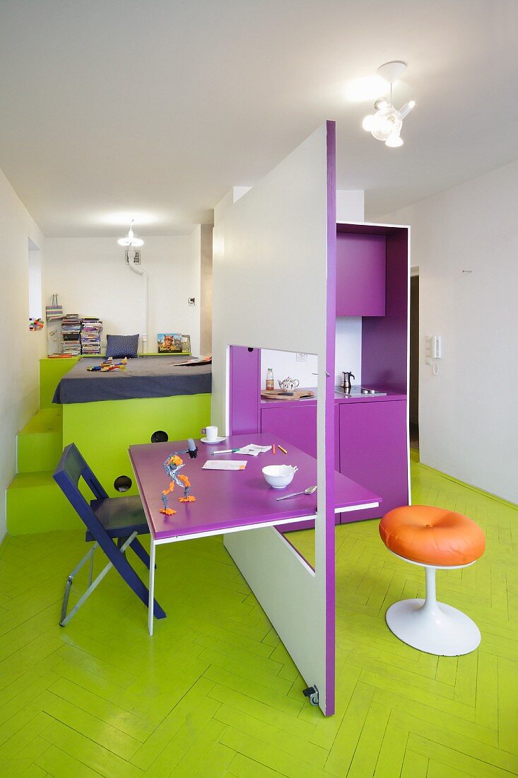 Modular kitchen with integrated dining table and bed on platform with storage space in colourful mini-apartment